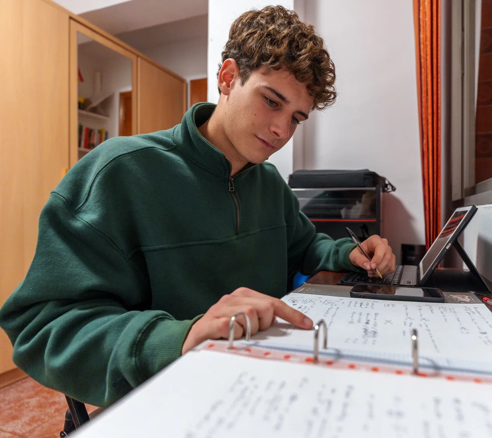 A high-school student working through handwritten math notes in a binder, with a tablet beside him